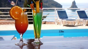 Dois cocktails com guarnições de frutas sobre uma mesa perto de uma piscina, com cadeiras de descanso e vista para o mar.
