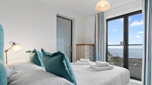 A bedroom with a large bed, a bedside lamp, a dresser, and a view of the sea through a large window.