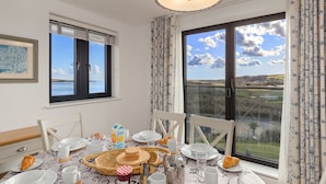 A dining area with a table set for breakfast, a view of the sea through large windows, and a ceiling light fixture.