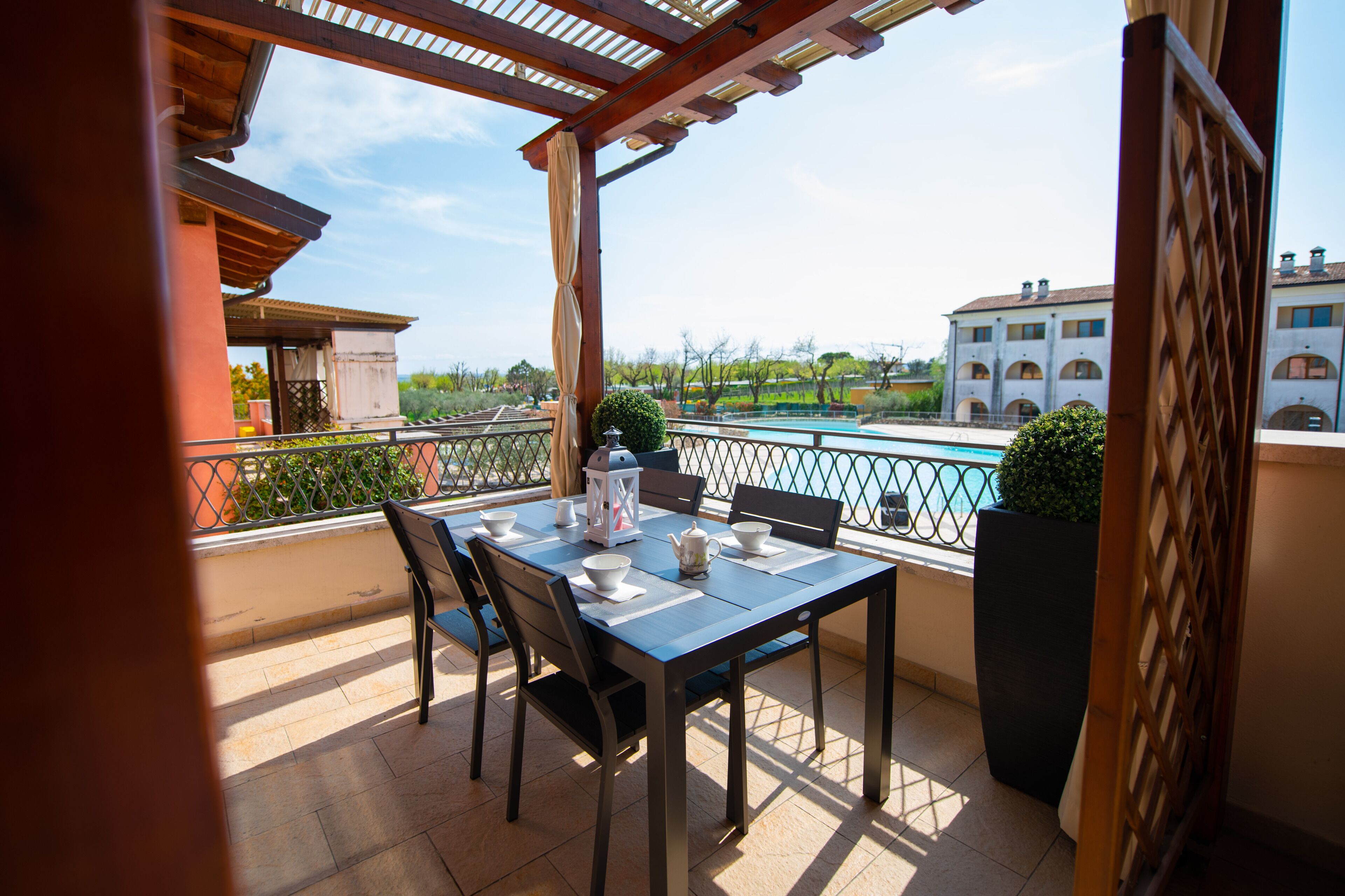 A balcony with a table set for two, chairs, a potted plant, and a view of a building and a pool.