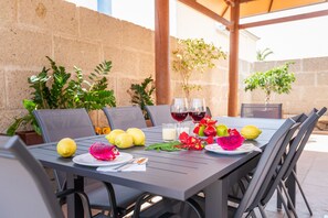 Un espacio exterior para comer con una mesa dispuesta para dos personas, que incluye varias frutas, dos copas de vino tinto y una vela.
