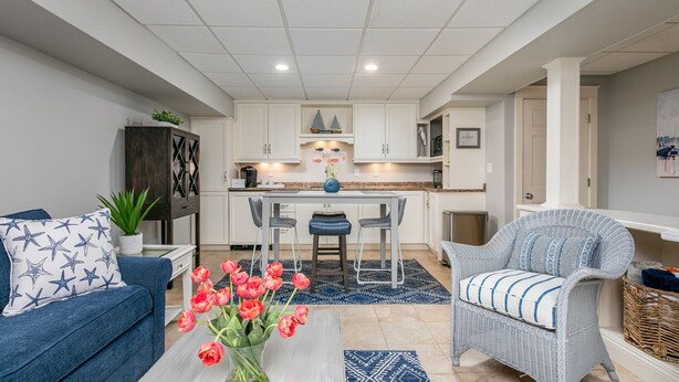 A modern living room with a sofa, a wicker chair, a coffee table with tulips, and a kitchen in the background. - Walk to Town, Riverfront Retreat & Beach Pass (Kennebunk)