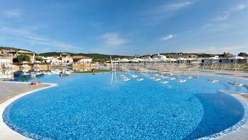 Ein großer, klarer blauer Pool mit Liegestühlen und Sonnenschirmen, umgeben von Gebäuden und Hügeln.