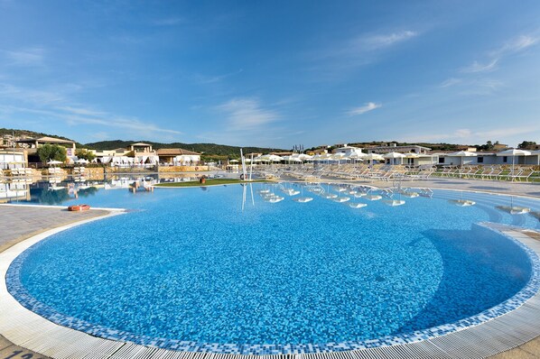 Une grande piscine d’un bleu éclatant, bordée de chaises longues et d’ombrelles, et entourée de bâtiments et de collines.