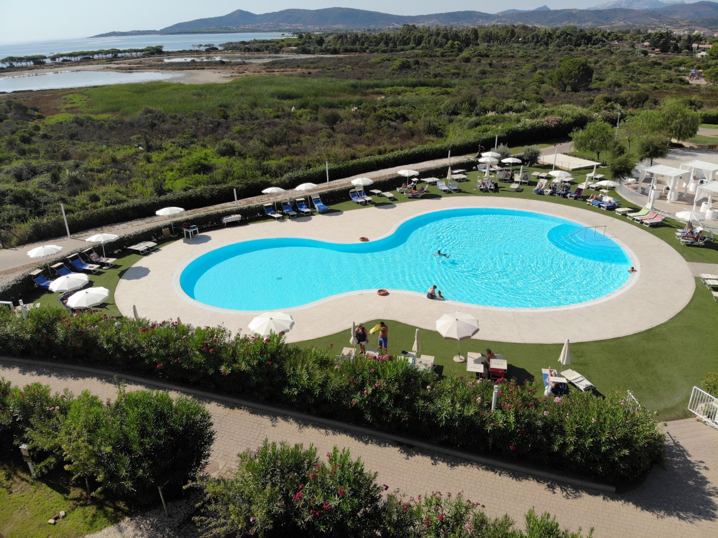 A large, circular swimming pool surrounded by lounge chairs and umbrellas, with a view of a natural landscape in the background.