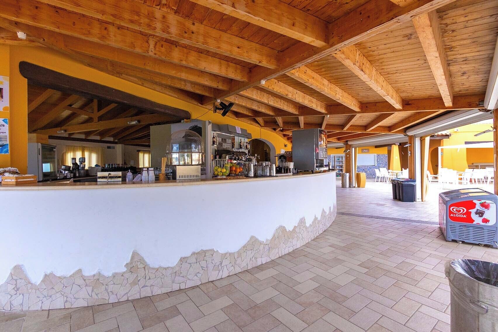 A covered outdoor area with a stone-tiled floor, a central counter with a coffee machine, and a trash bin labeled 'ALGIDA'.