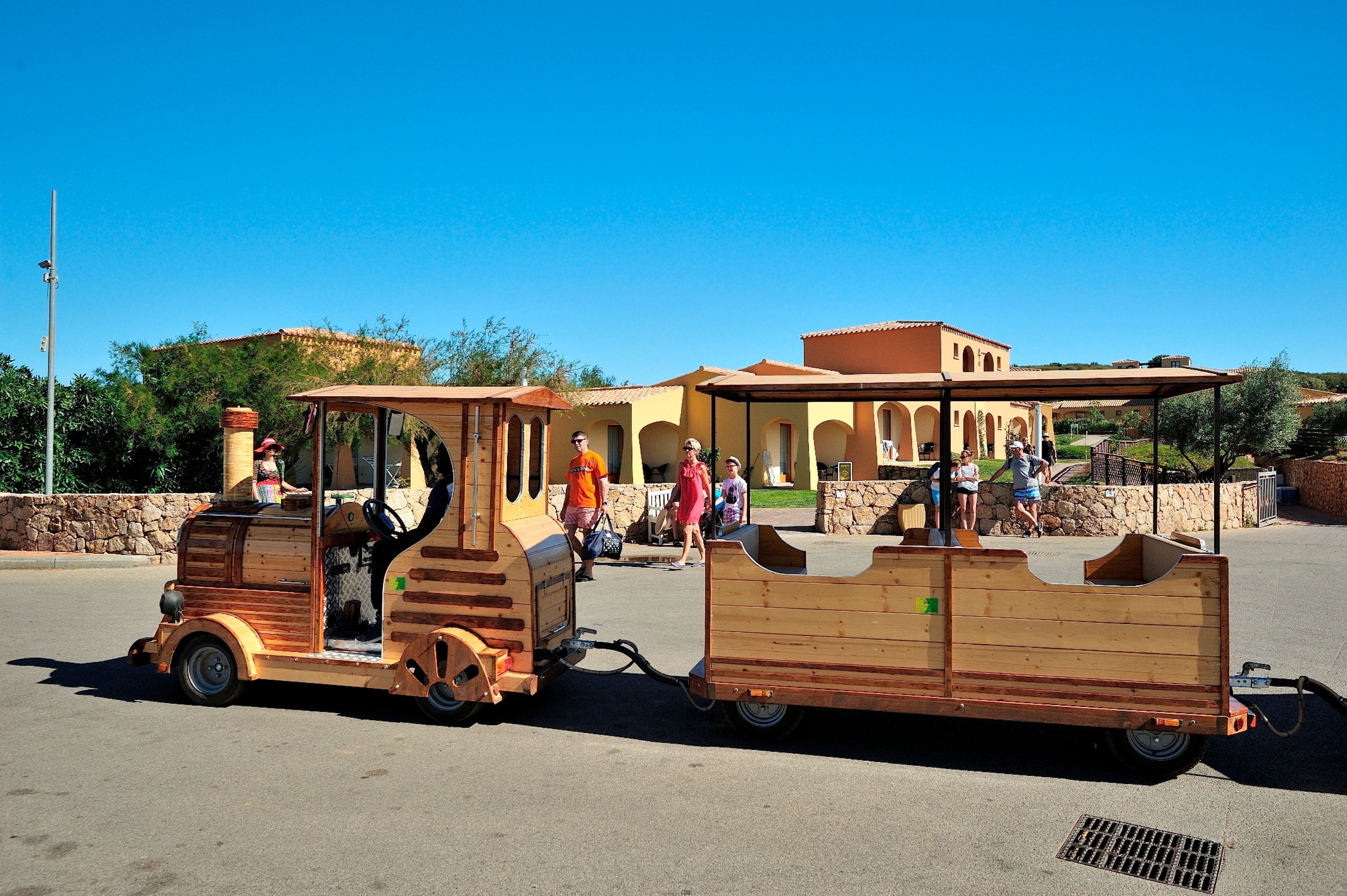Un treno di legno antico con passeggeri, ambientato in una zona all'aperto soleggiata con pareti in pietra e cielo azzurro.