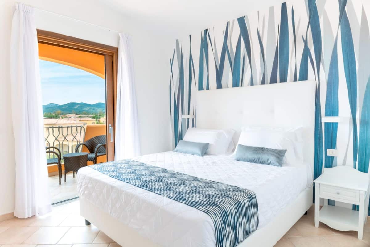 A hotel room with a large bed, a white nightstand, a view of the outdoors through a balcony, and a patterned wall.