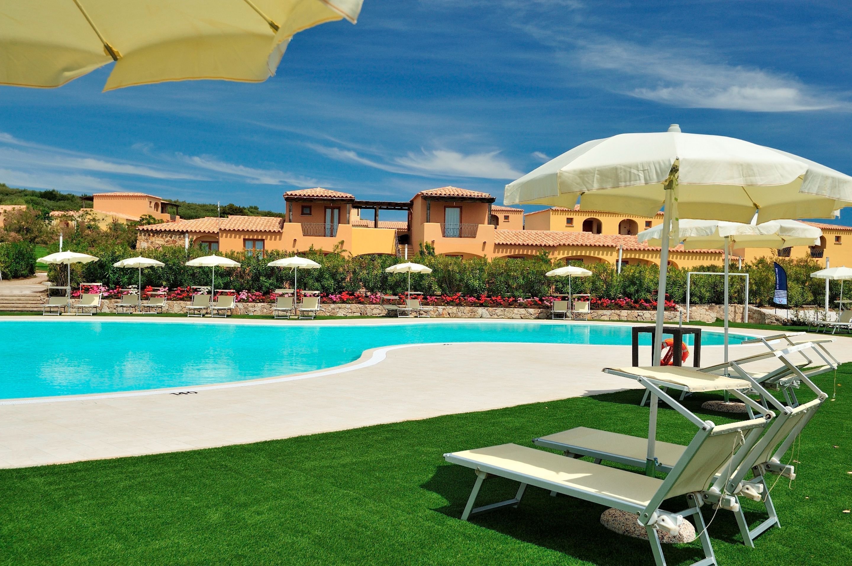 Una piscina con sillas de descanso y sombrillas, rodeada de un césped bien cuidado y edificios con techos de teja.