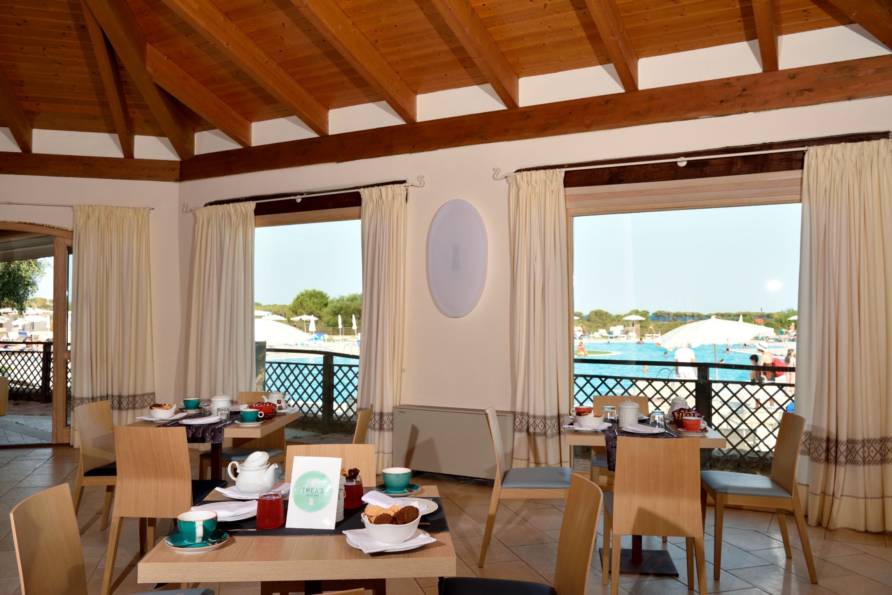 Un espace repas avec des tables et des chaises en bois, une vue sur une piscine et un balcon doté d’une balustrade.