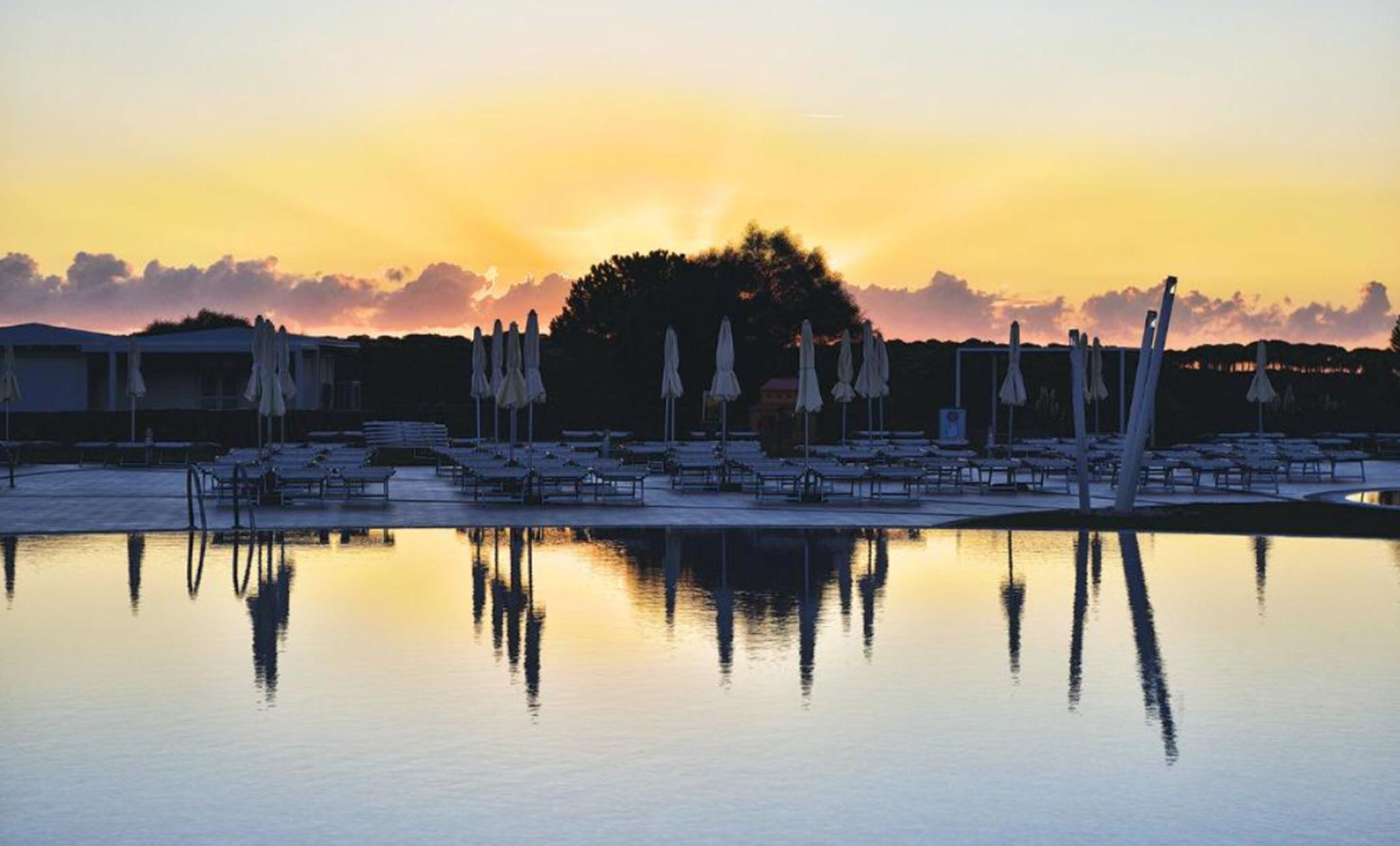 A poolside area with lounge chairs and umbrellas, reflecting in the water during sunset.