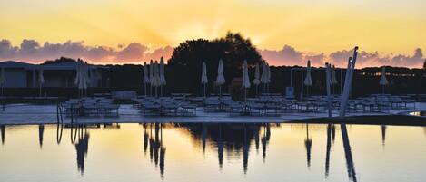 A poolside area with lounge chairs and umbrellas, reflecting in the water during sunset.