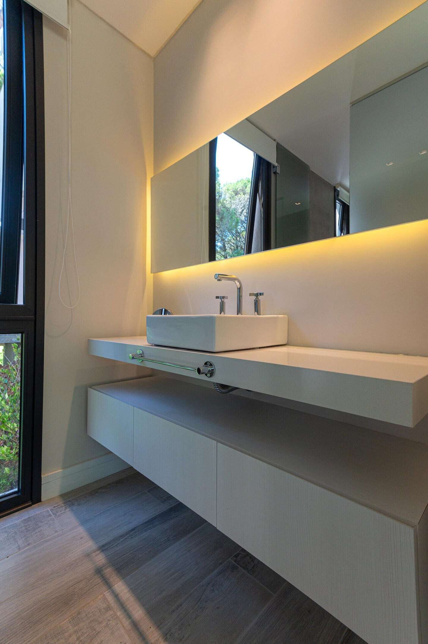 A modern bathroom with a large mirror, a white sink, and a wooden floor.