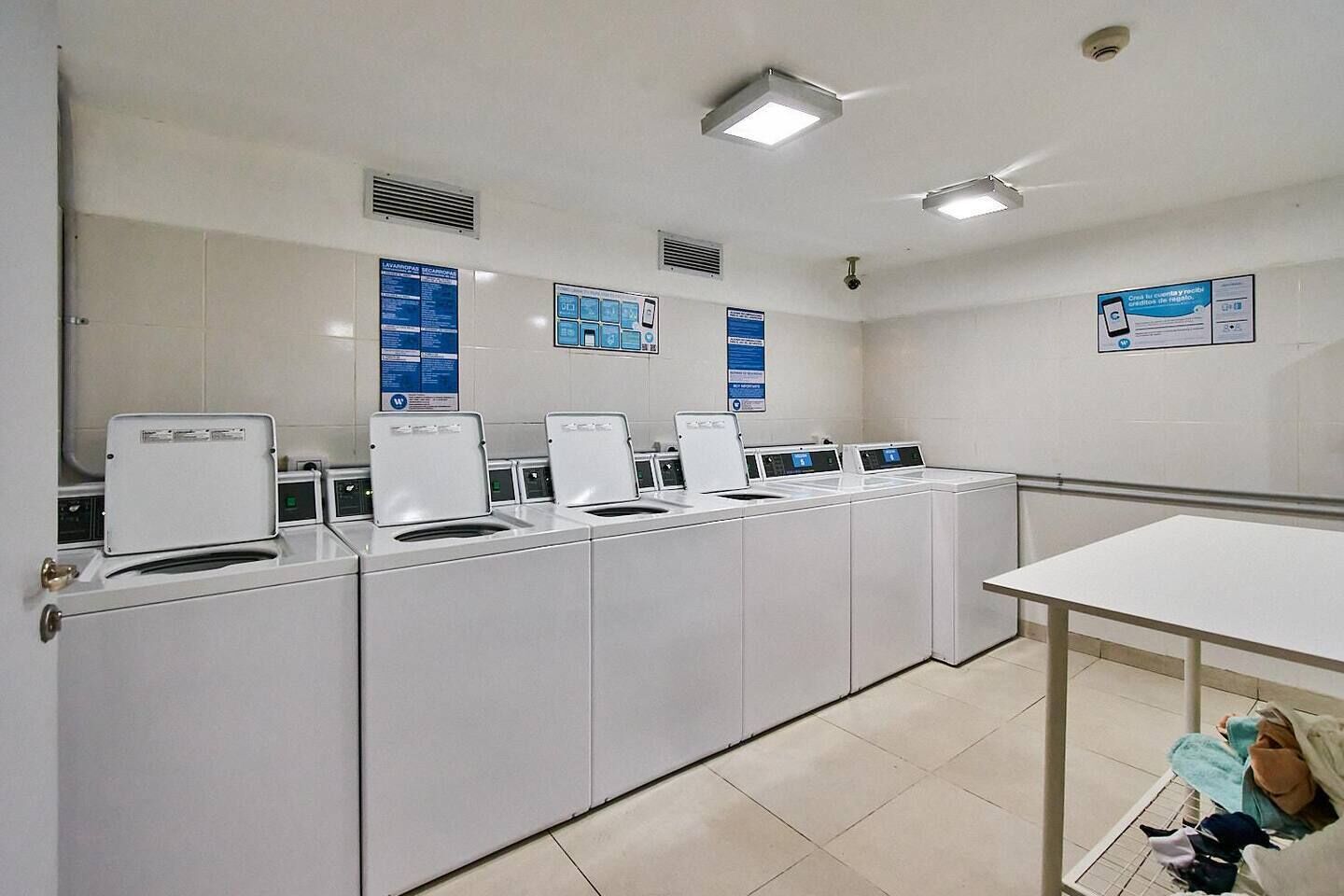 A laundry facility with multiple washing machines and dryers, a tiled floor, and informational posters on the walls.