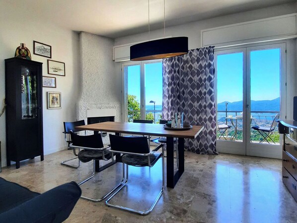 A modern living room with a dining area, large windows with a view, and a black cabinet with framed pictures.