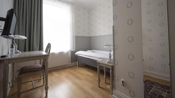 A bedroom with a single bed, a bedside table, a chair, a desk with a lamp, and a window with curtains.