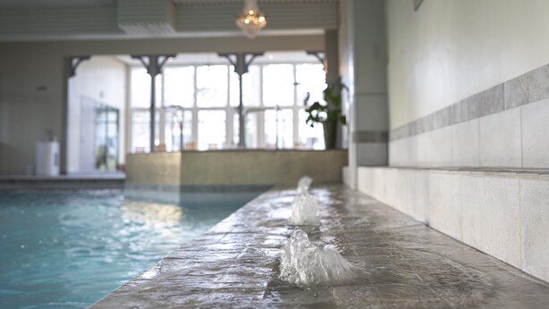Una piscina interna con acqua limpida, un solarium in legno e vista su un edificio con ampie finestre e un lampadario.