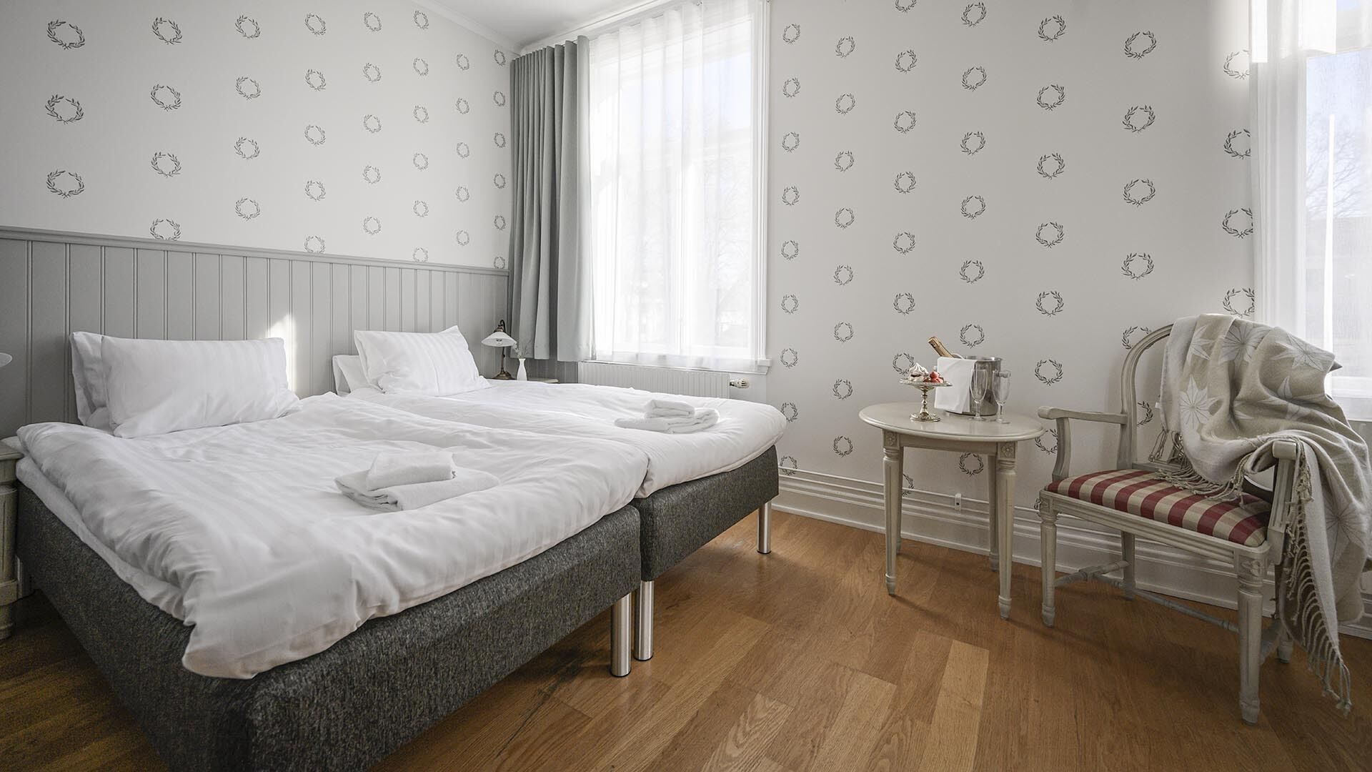 A neatly made bed with white linens, a grey headboard, and a small round table with a bottle and glasses.