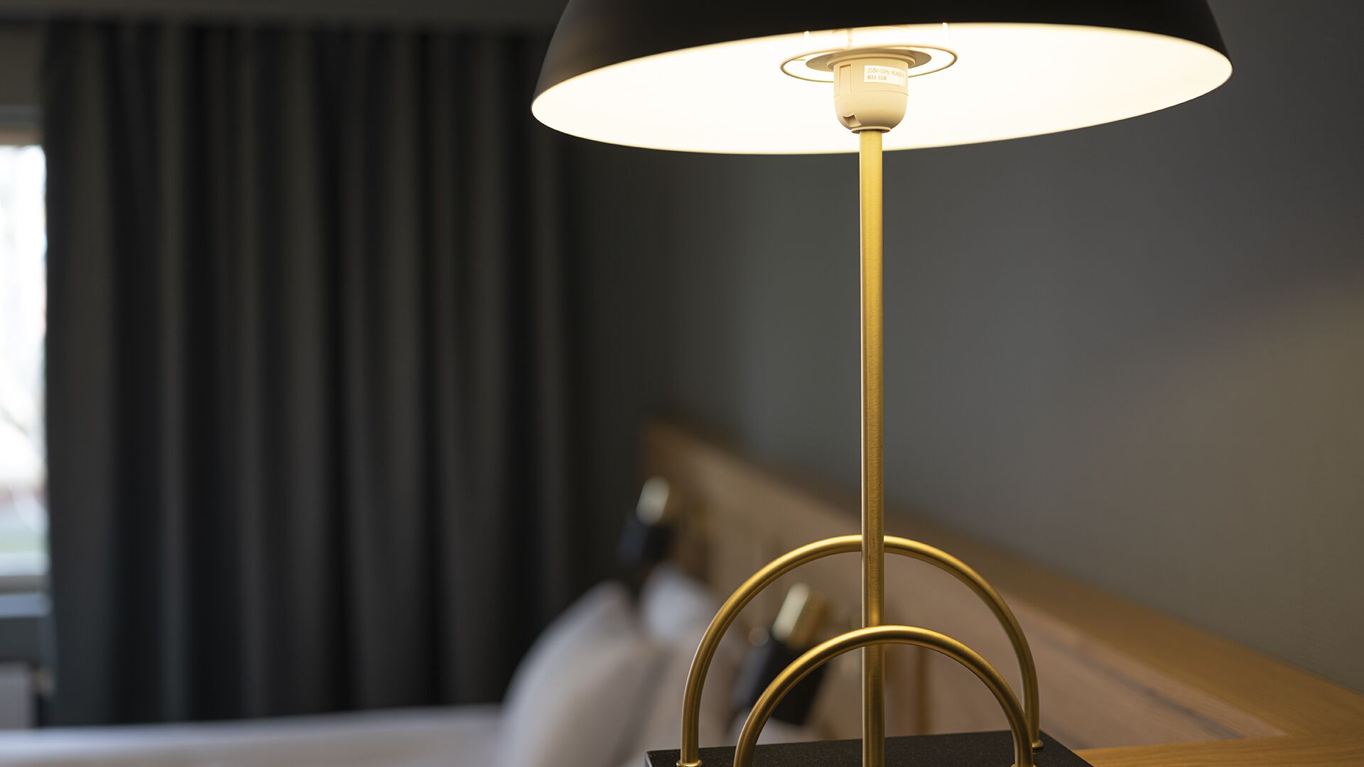 A modern floor lamp with a black shade and a gold base, positioned on a wooden table in a room with a dark curtain and a bed in the background.