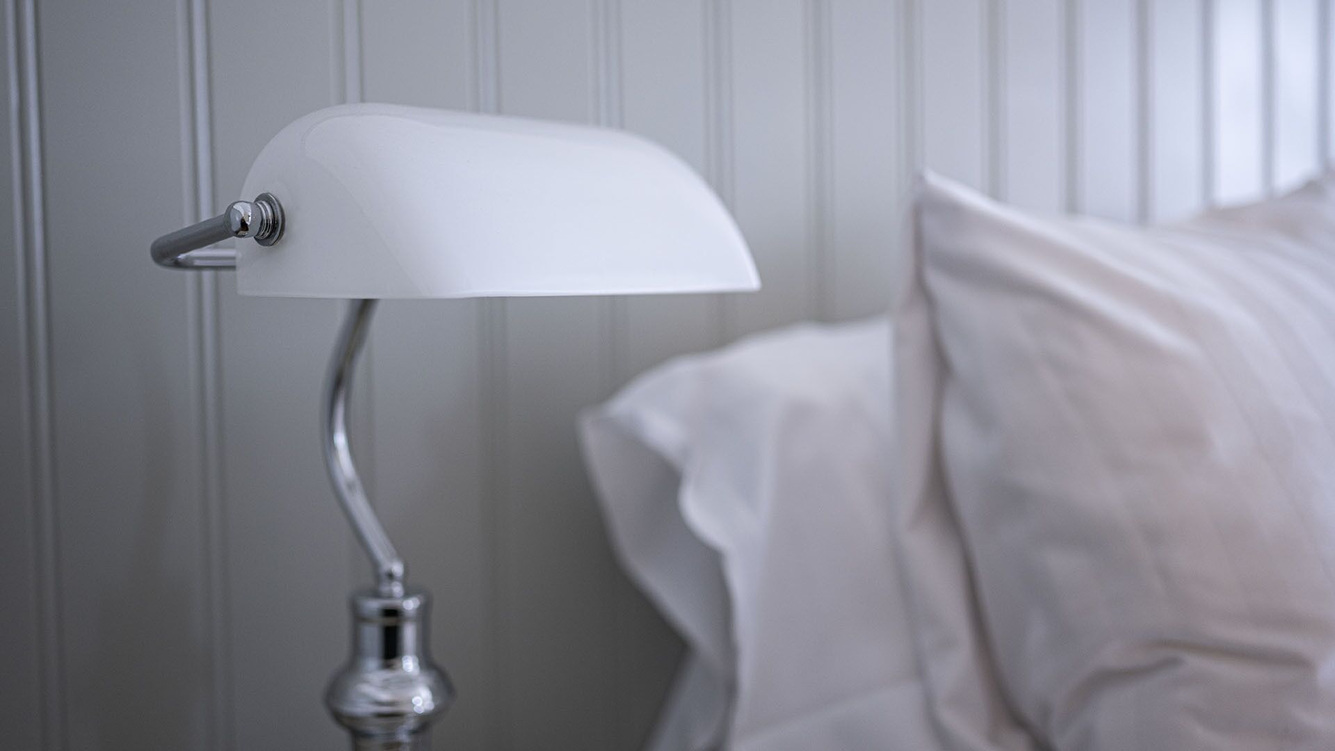 Una lámpara de noche blanca con brazo curvo y base metálica, ubicada junto a una cama con ropa blanca de cama.