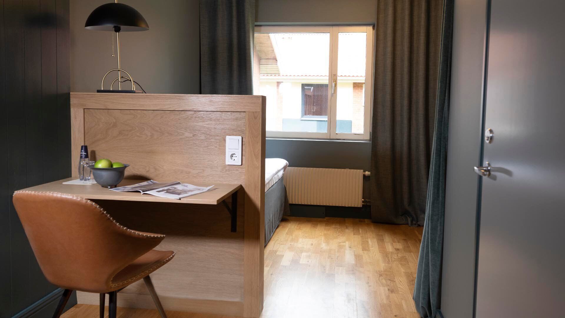 A compact room with a wooden desk, a chair, a radiator, a window with curtains, and a door.