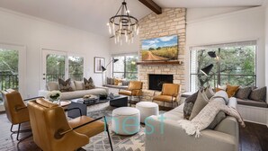 A spacious living room with a stone fireplace, leather furniture, and large windows offering views of the outdoors. - ***NEW*** 7 MINUTES TO DOWNTOWN | POOL | HOT TUB | POOL TABLE | FOOSBALL | FIRE PIT | 13 Queen Beds (Austin)