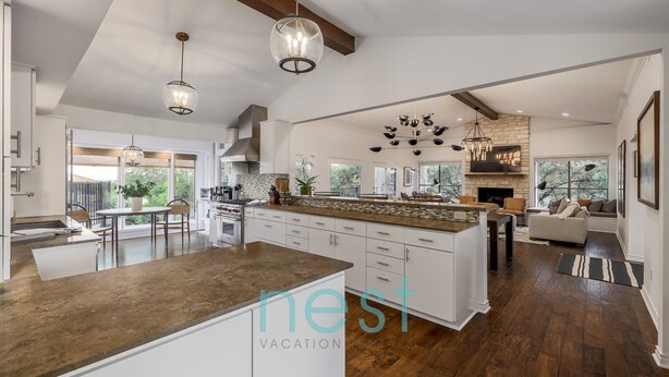 A spacious kitchen with a large island, white cabinetry, and a stone backsplash. There is a dining area with a table and chairs, and a living room with a sofa and a fireplace. - ***NEW*** 7 MINUTES TO DOWNTOWN | POOL | HOT TUB | POOL TABLE | FOOSBALL | FIRE PIT | 13 Queen Beds (Austin)