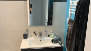 Un baño con lavamanos blanco, tocador negro y un espejo. Hay artículos de aseo en el lavamanos, un perchero y una cesta debajo del tocador.