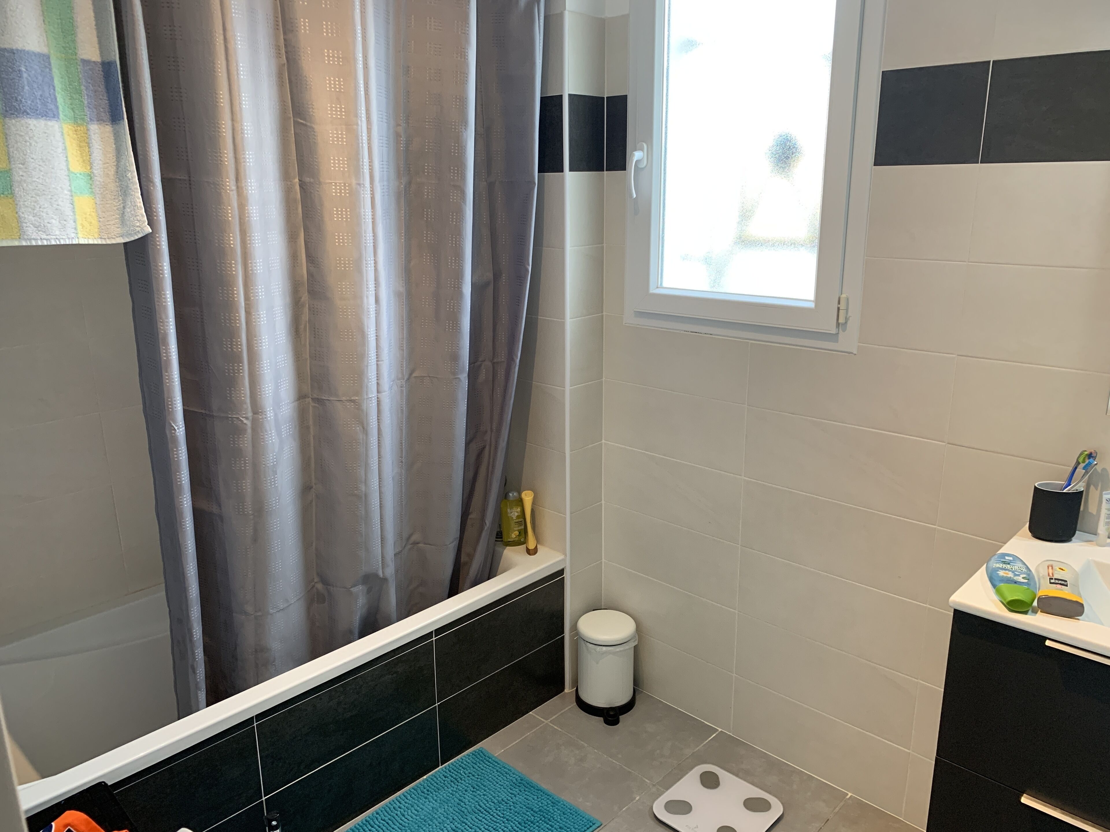 A bathroom with a shower curtain, a toilet, a waste bin, a scale, and toiletries on the counter.