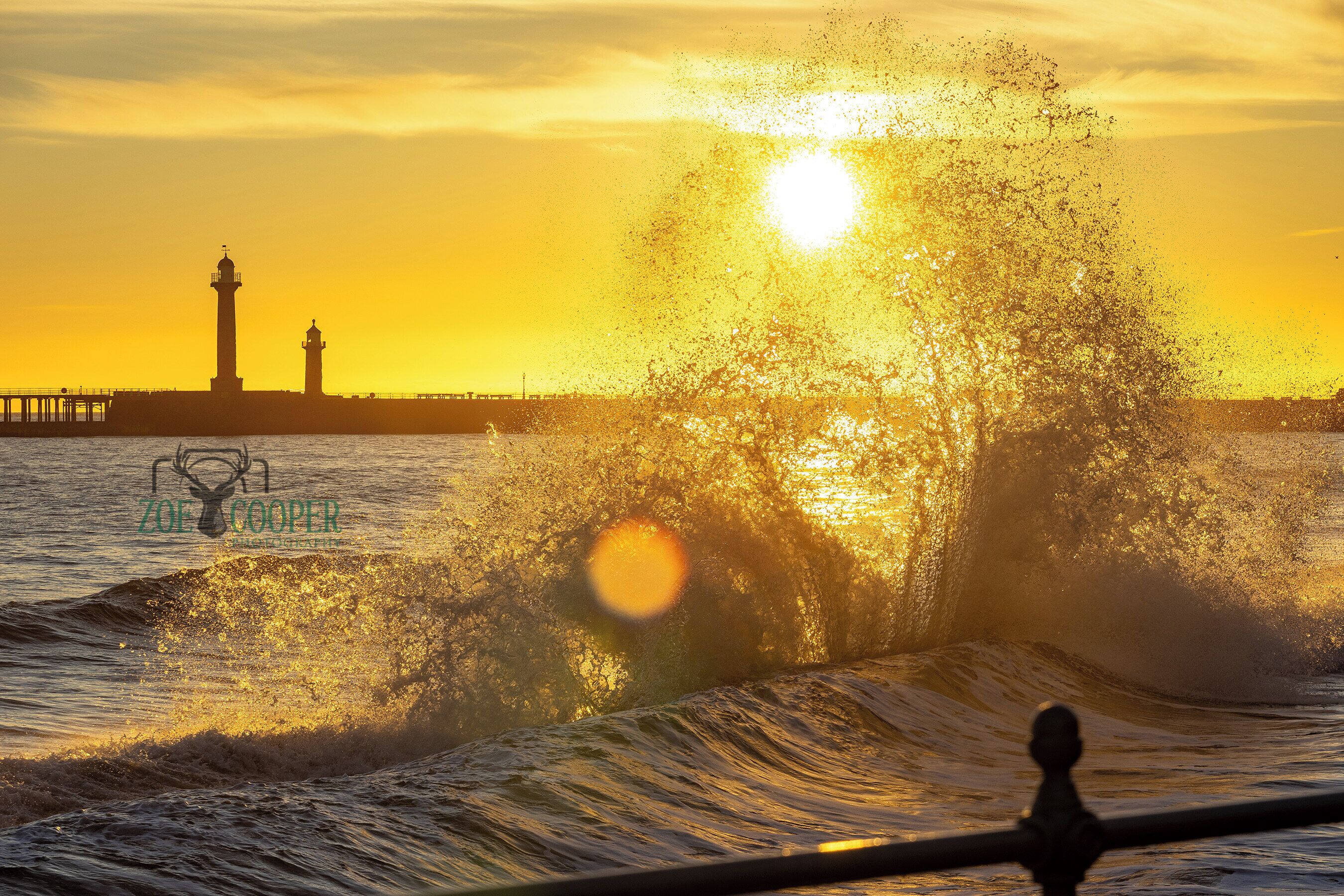 Silhouet van een vuurtoren tegen een zonsondergang, met golven die tegen de kust slaan, en een pier op de achtergrond.