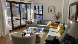 Un salon moderne avec un canapé, des fauteuils et une table basse. De grandes fenêtres offrent une vue sur l’extérieur.