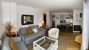 Un salon moderne avec un canapé gris, un fauteuil blanc et une table basse en verre. On aperçoit, au fond, une cuisine avec un coin repas.