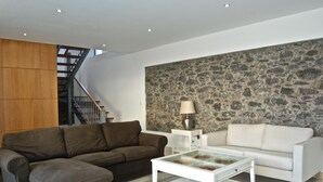 A modern living room with a brown sectional sofa, a white sofa, and a glass coffee table. There is a stone wall and a wooden panel on the left side. - Casa da Praia Calheta - Moradia (Calheta)