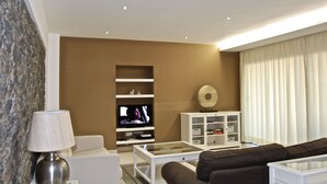 A modern living room with a flat-screen TV, a glass-top coffee table, a brown sofa, and a white cabinet with shelves. - Casa da Praia Calheta - Moradia (Calheta)