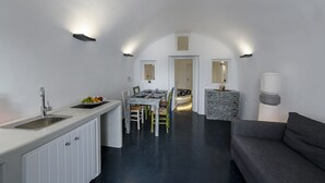 Une cuisine moderne avec des armoires blanches, un évier en acier inoxydable et un panier à fruits. On y trouve un coin repas avec une table et des chaises, ainsi qu’un canapé gris dans le salon.