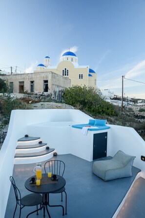 A sunlit outdoor seating area with a table and chairs, a cushioned bench, and a view of a building with blue domes. - The Luna Cavern 1 Sky Blue (Santorini)
