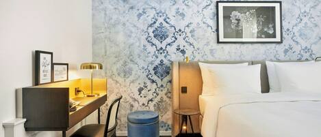 A hotel room with a bed, a desk with a lamp, a chair, a small stool, and a framed picture on the wall.