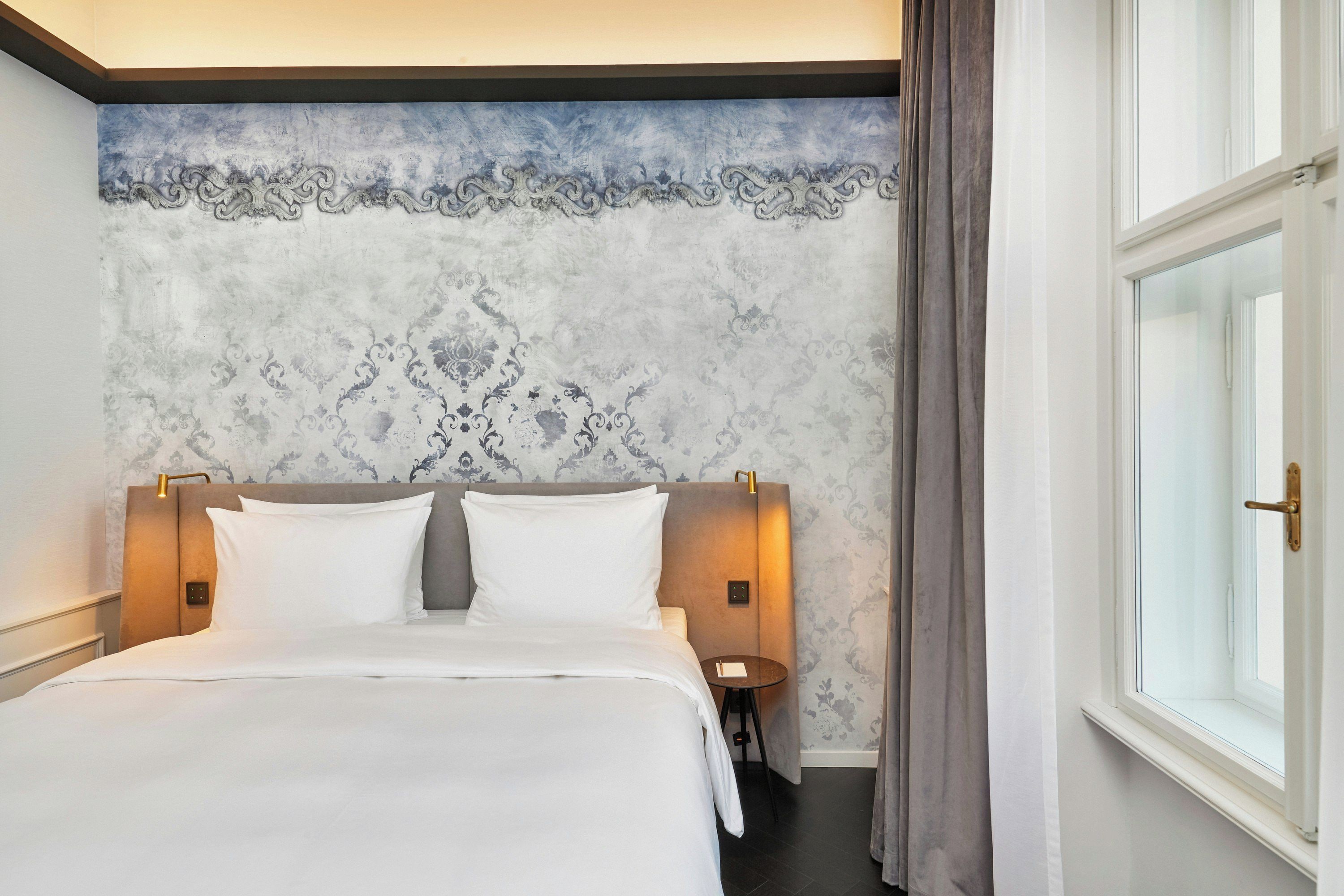 Una habitación de hotel con una cama grande, un cabecero con lámparas fijadas a la pared, una mesita y una ventana con cortinas.