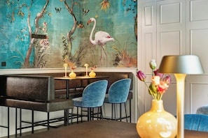 A dining area with a mural, two blue chairs, a table with candles, and a vase with flowers.