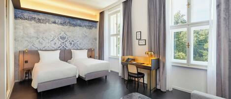 A hotel room with two beds, a desk, a chair, a sofa, and large windows with curtains.