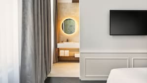 A modern hotel room with a large bed, a flat-screen TV mounted on the wall, a round mirror with a light, and a bathroom with a sink and towel rack.