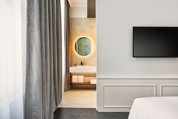 A modern hotel room with a large bed, a flat-screen TV mounted on the wall, a round mirror with a light, and a bathroom with a sink and towel rack. - The Cloud One Salzburg (Salzburg)