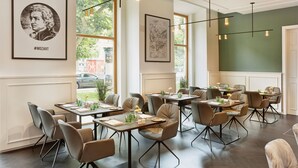 A restaurant with multiple tables and chairs, framed artwork on the walls, and large windows allowing natural light.