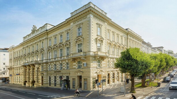 Un edificio grande e histórico con arquitectura clásica, situado en una esquina de calle con un árbol y coches estacionados.