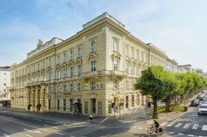 Un grande edificio storico dall'architettura classica, situato all'angolo di una strada con un albero e auto parcheggiate.