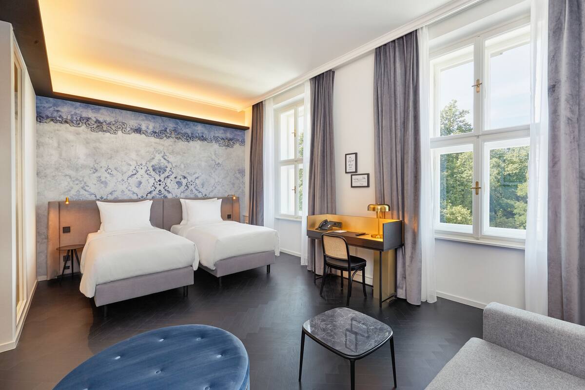 Habitación de hotel con dos camas, un escritorio, una silla, un sofá y un ventanal con cortinas.