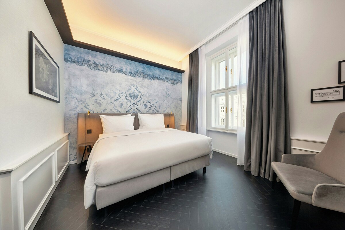 Habitación de hotel con una cama grande, una silla, una ventana con cortinas y una pared con un diseño decorativo.