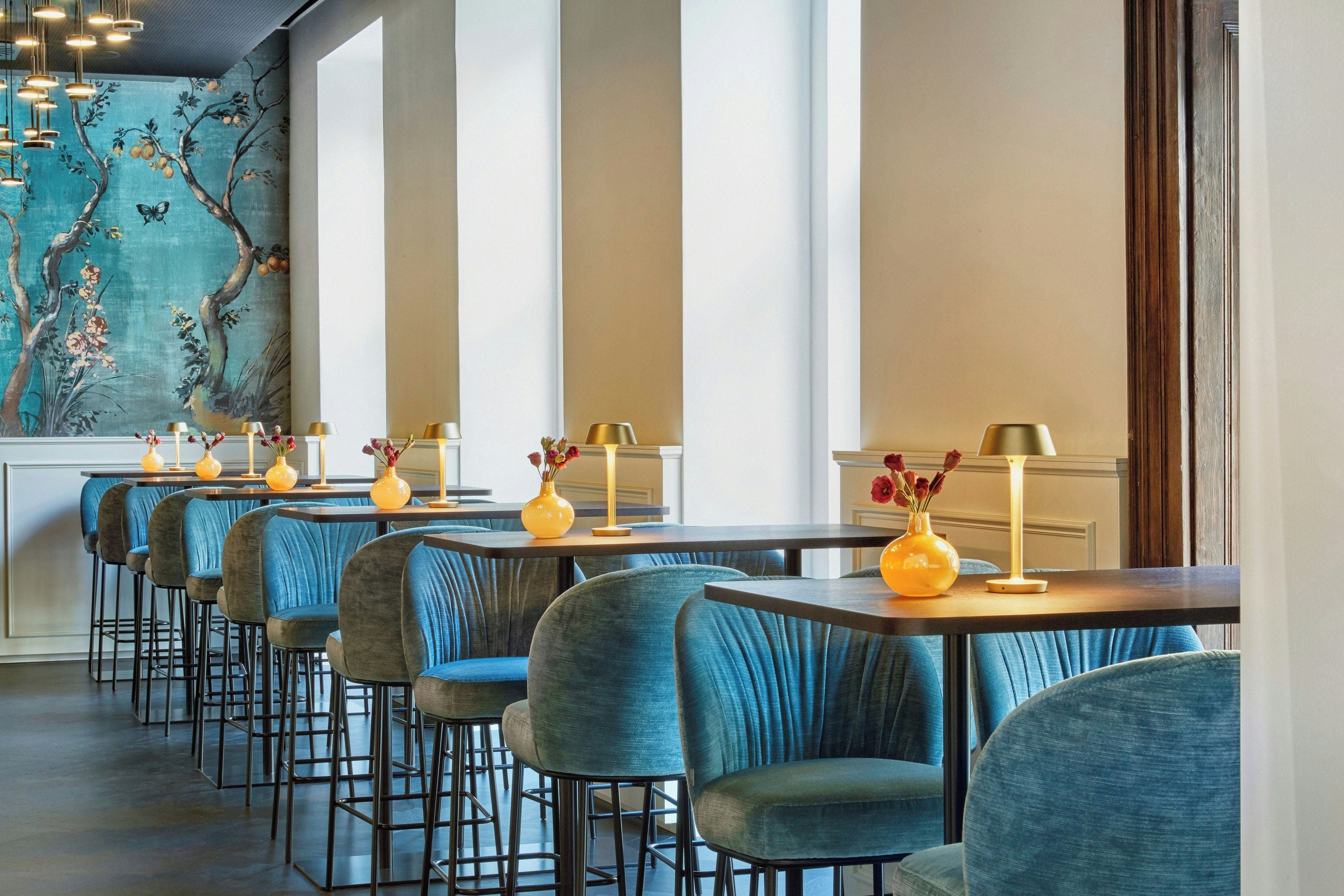 A restaurant interior with high tables and blue upholstered chairs, decorated with yellow vases and lamps.
