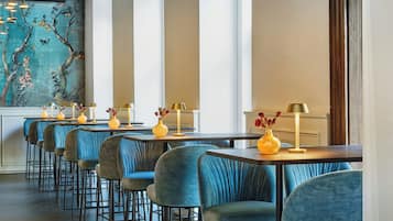 A restaurant interior with high tables and blue upholstered chairs, decorated with yellow vases and lamps.