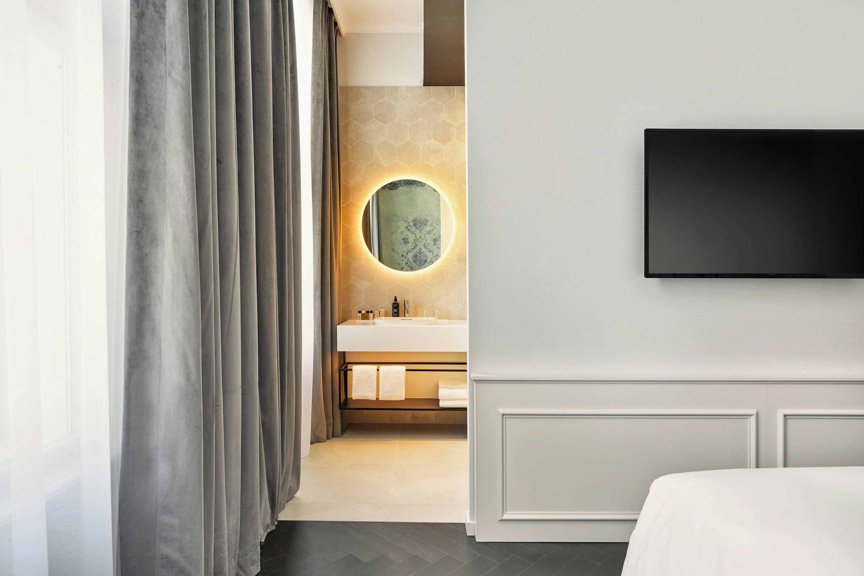 Ett modernt hotellrum med en stor säng, en platt-TV som är monterad på väggen, en rund spegel med en hexagonformad ram och ett badrum med en vit handfat och handduksställ.